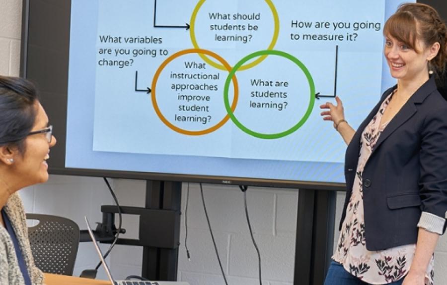 Natasha Holmes standing in front of a graphic that asks "What should students be learning? What instructional approaches improve student learning? What are students learning? What variables are you going to change? How are you going to measure it?"